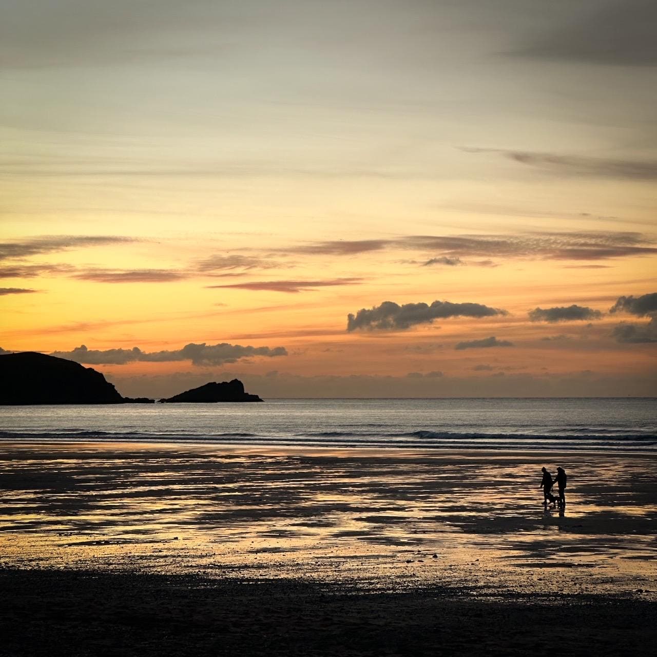 Two people walking on a beach at sunset with a silhouetted headland in the background