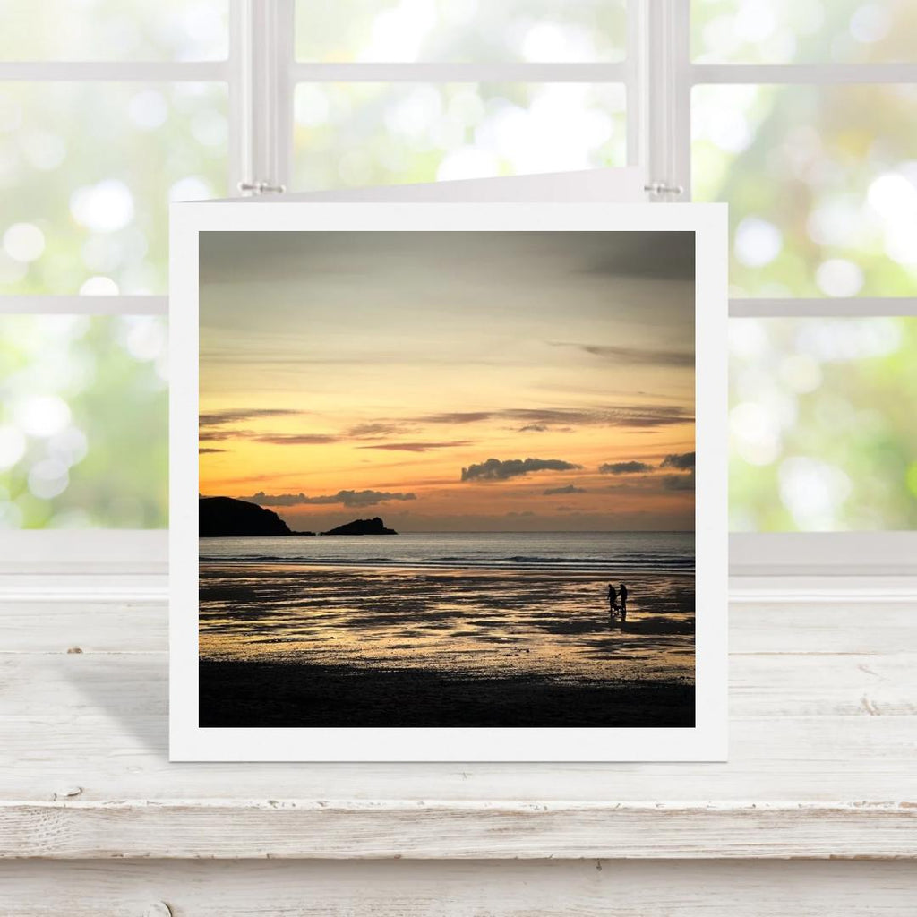 Square greetings card with photograph of a sunset over a beach with two people standing on the shore, displayed on a white surface.