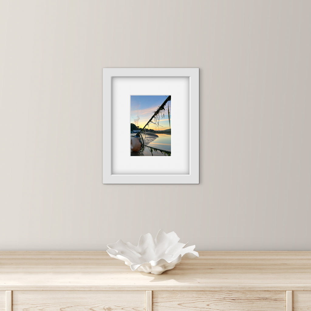 Framed artwork of a sunrise reflected over water with mooring ropes and a bouy on a wall above a wooden surface with a decorative bowl.
