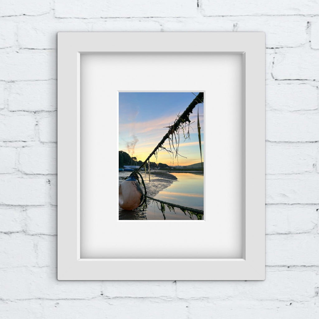 Framed photograph of a sunrise over water with mooring ropes on a river in a white frame on a white brick wall.