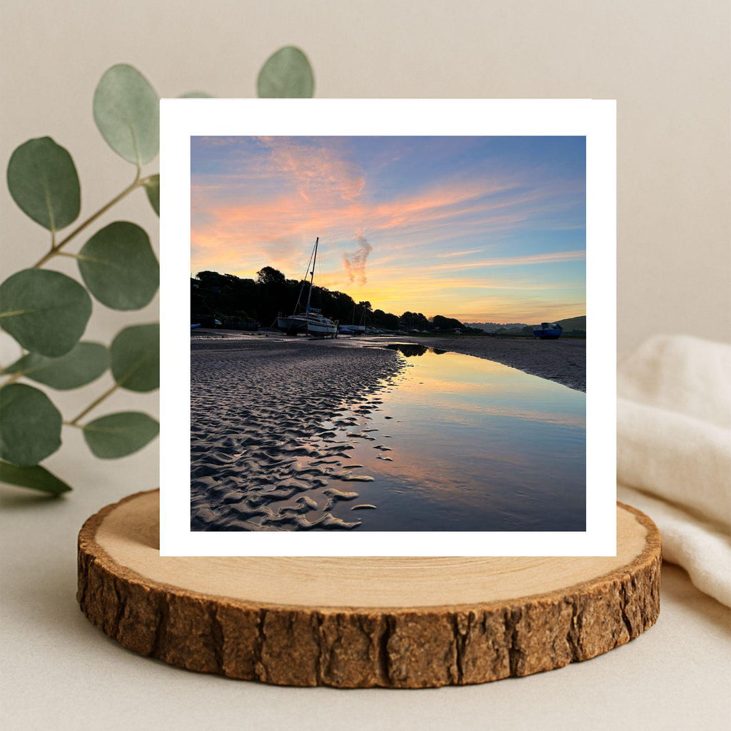 Greetings card of a sunset reflected  in water . Displayed on a wooden stand with a plant and blanket in the background.
