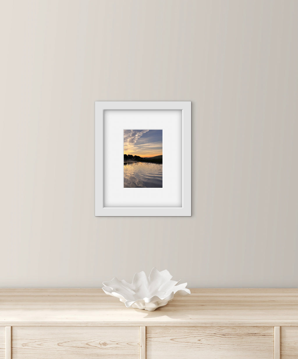 Framed photograph of a sunrise over water on a wall above a wooden cabinet with a decorative bowl.