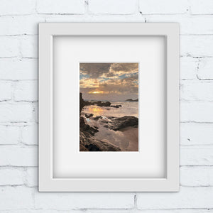 Framed photograph of a beach scene with a rocky shore with sunset on a white brick wall.