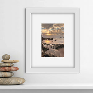 Framed beach sunset scene print on a white wall with stacked stones below