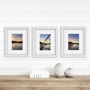 Three framed photographs of river landscapes with a pastel sunrise on a wall above a wooden shelf with decorative vases.