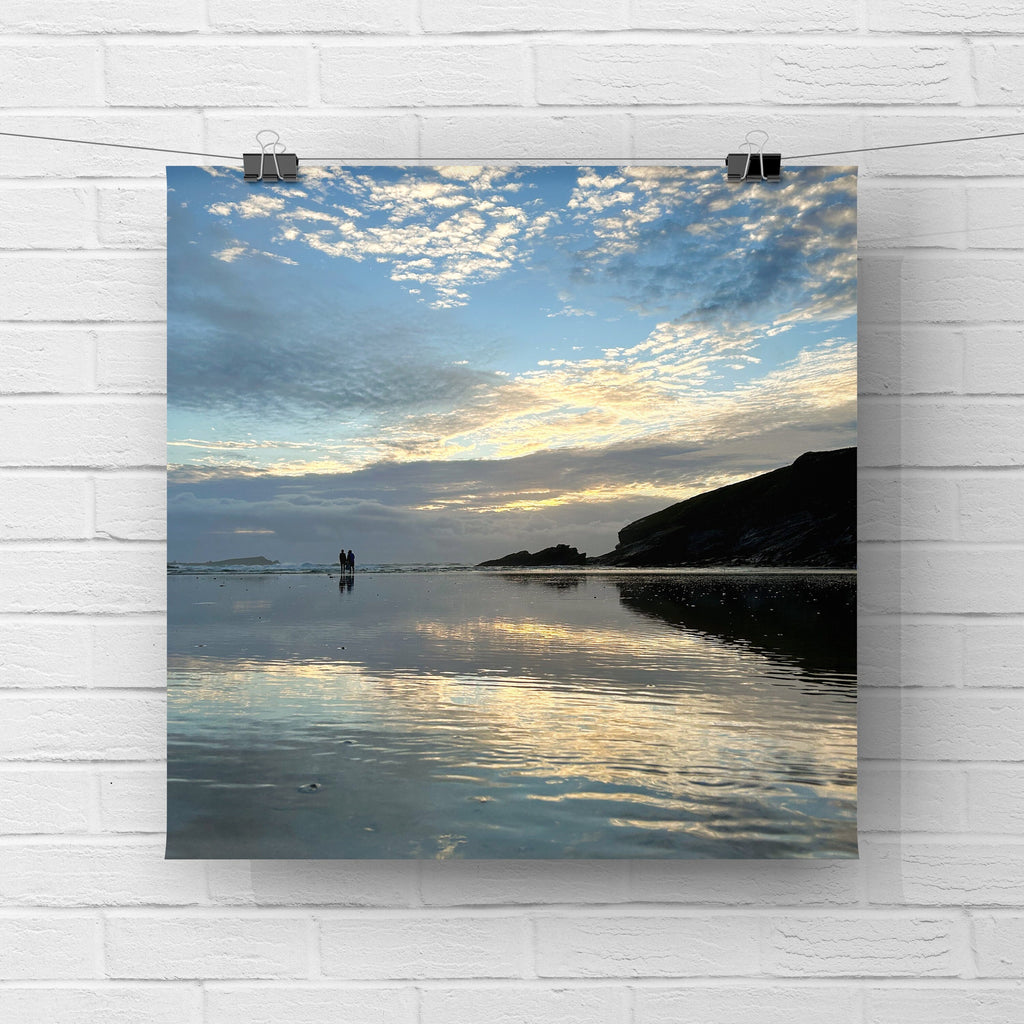 Poster of a beach scene with people standing on wet sand and a sunset reflection. The print is  hanging on a white brick wall.