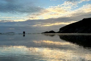 Two people standing on a beach with a reflective water surface and a dramatic sunset sky.