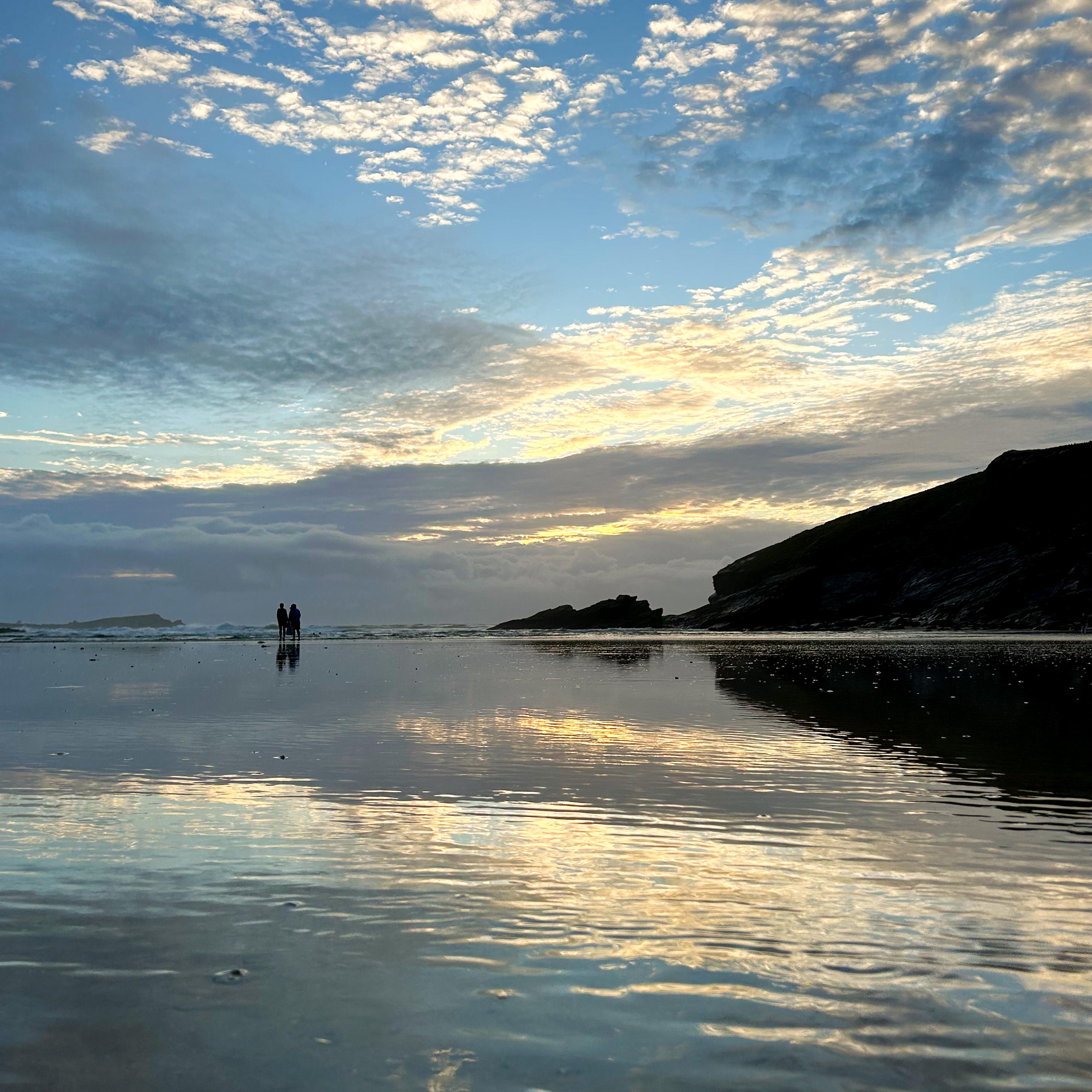 Two people standing on a beach with a reflective water surface and a dramatic sky.
