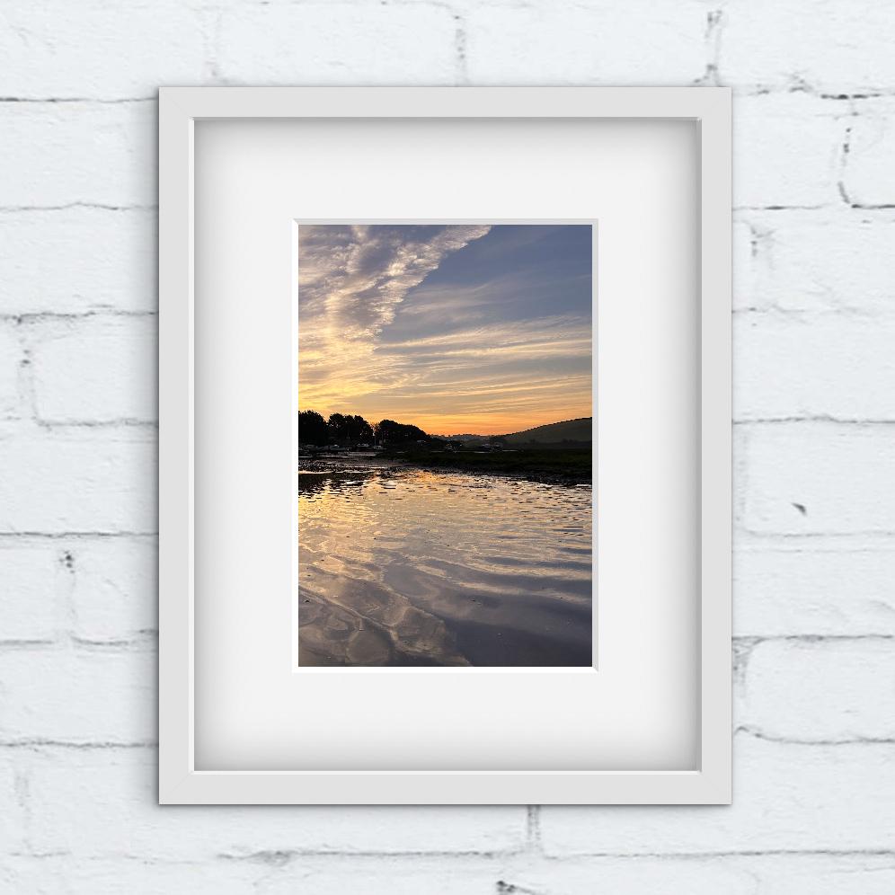 Framed photograph of a sunrise over water on a white brick wall.