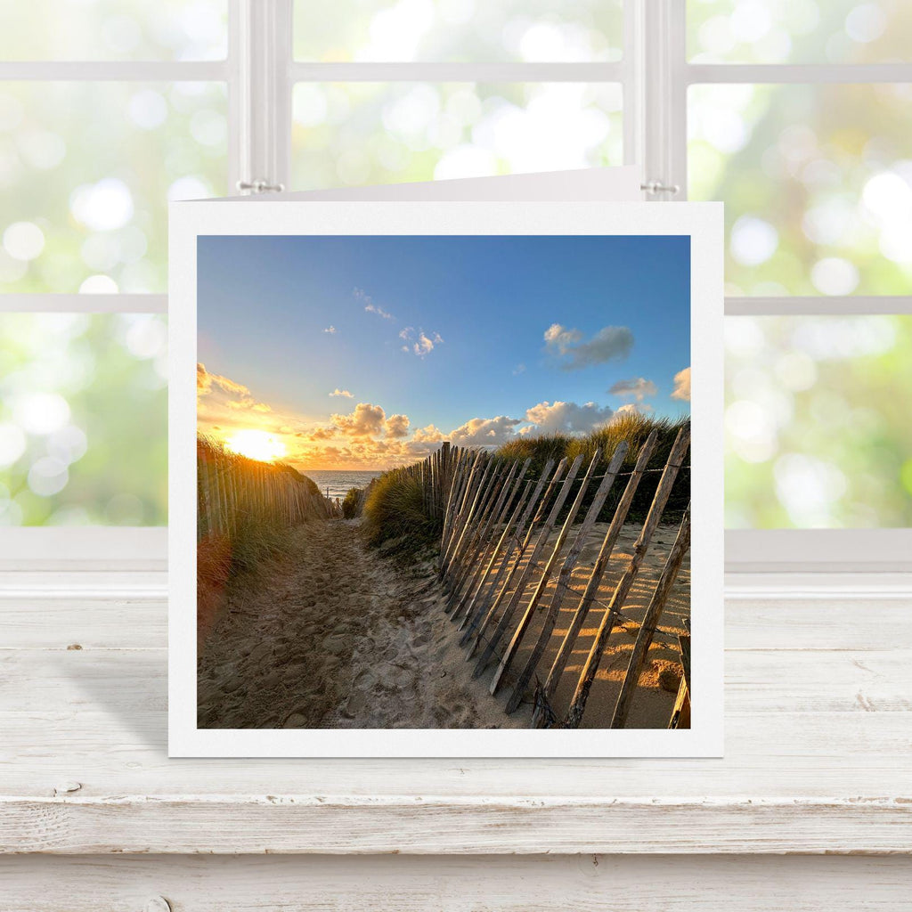 Square greetings card with photograph of a beach scene with a sunset over the ocean, displayed on a white surface.