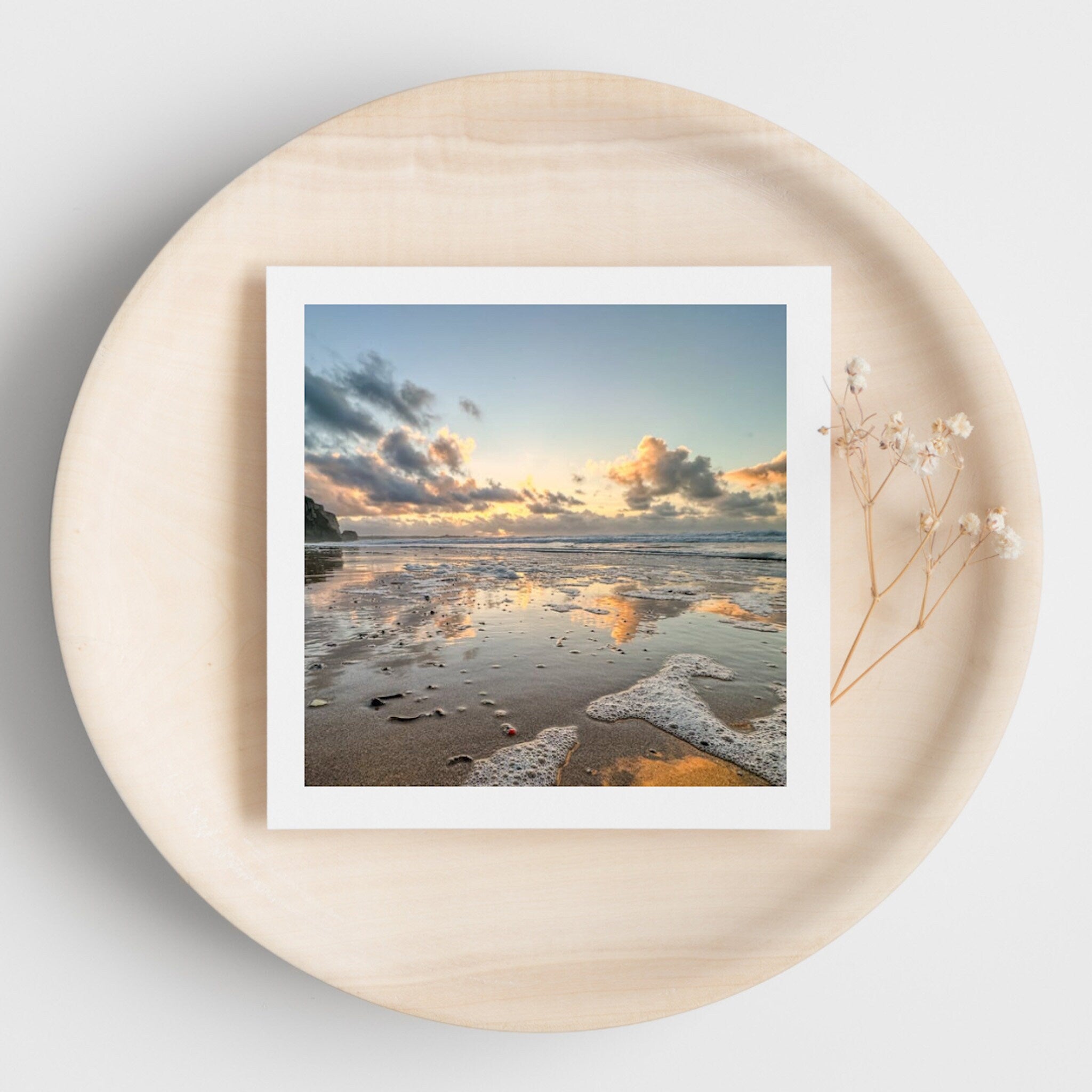 White edged beach sunset greetings card on a round beige surface with dried flowers.