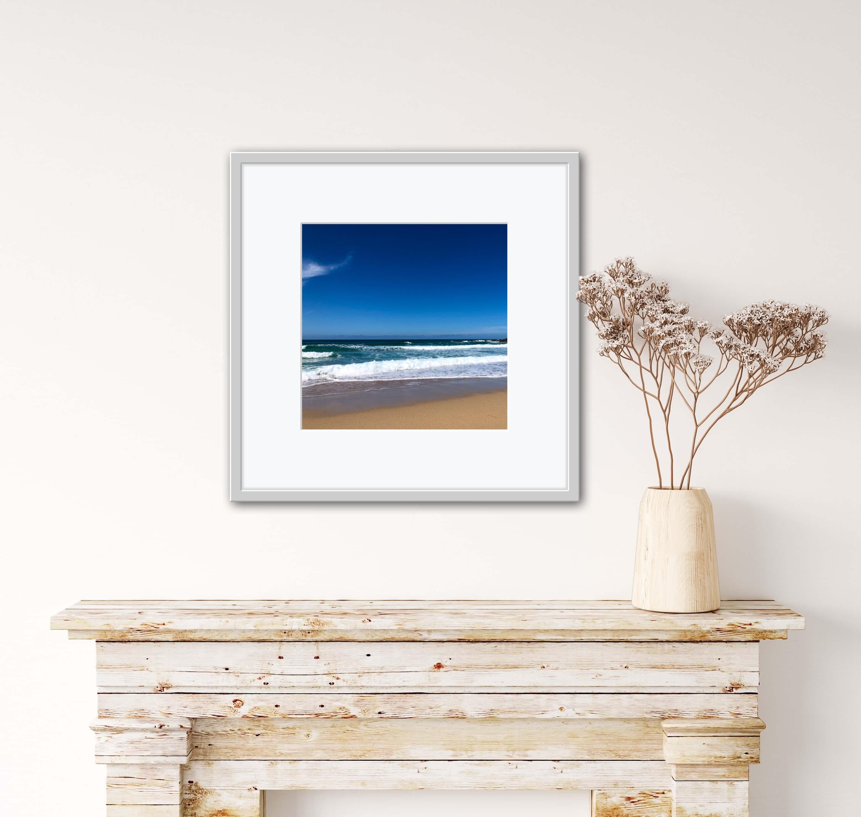 Framed beach print on a wall above a wooden console table with a vase of dried flowers.