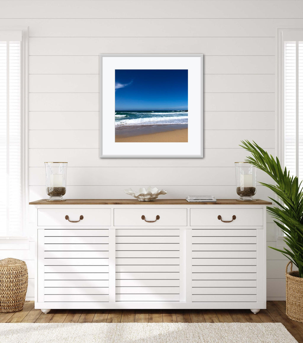 Framed beach scene art on a wall above a white cabinet with wooden top.