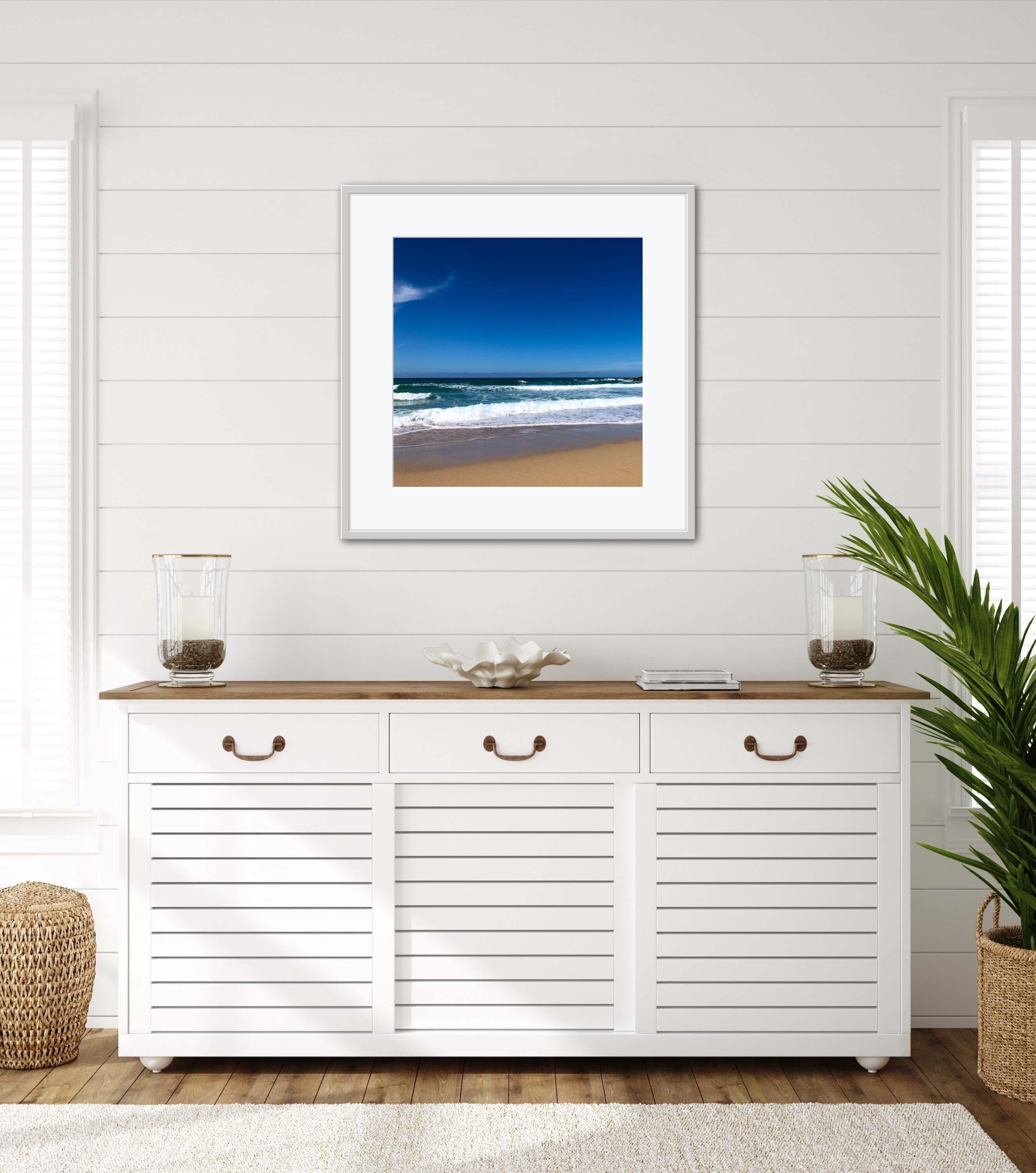 Framed beach scene art on a wall above a white cabinet with wooden top.