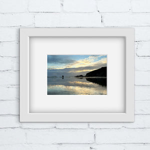 Framed photograph of a blue and gold coastal scene with a sunset over water on a white brick wall.