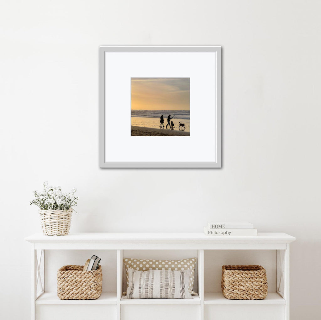 Framed photograph of a family with a dog on a beach at sunset, displayed on a wall above a white shelf.